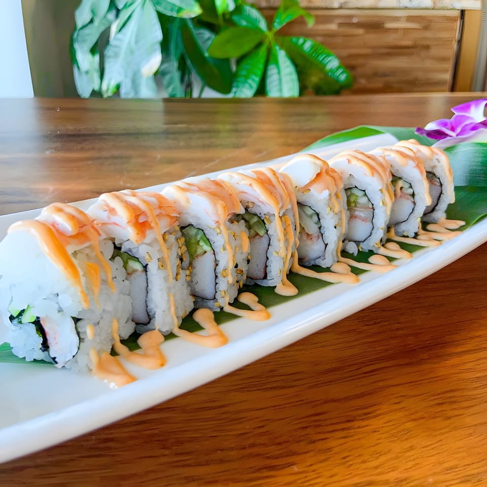Sushi roll with white rice, green filling, topped with shrimp and orange sauce, on a white plate.
