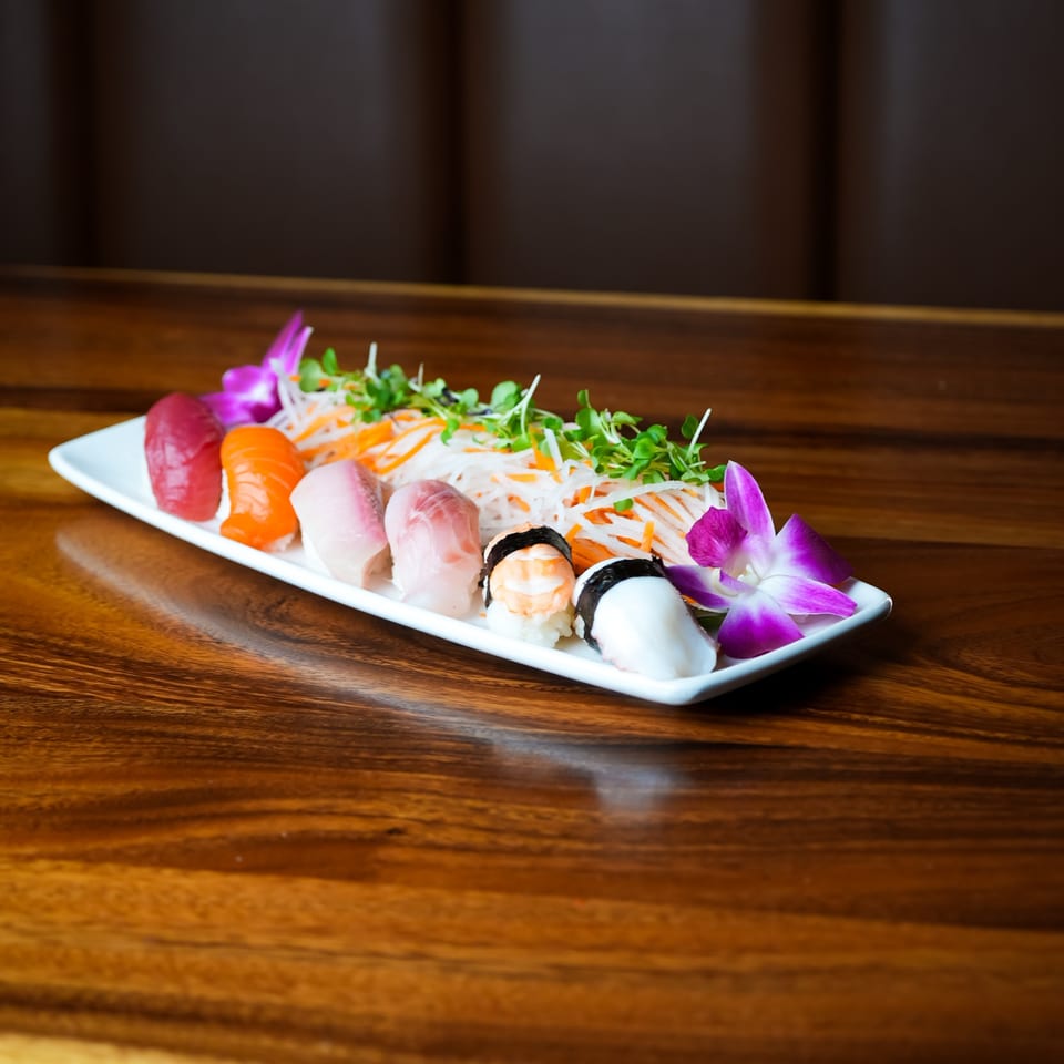 A white rectangular plate of various sushi and sashimi, garnished with microgreens and purple orchids.