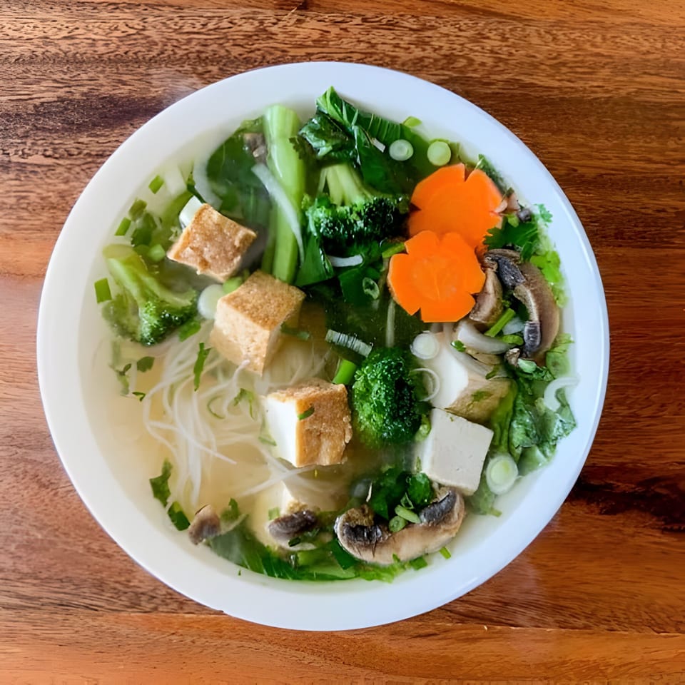 A white bowl of Koy Restaurant's pho with noodles, tofu, broccoli, carrots, and mushrooms.